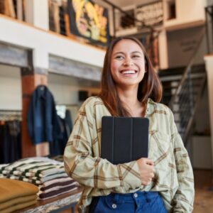 una joven sonriente sosteniendo una tableta digital de pie frente a un exhibidor de ropa en una tienda minorista