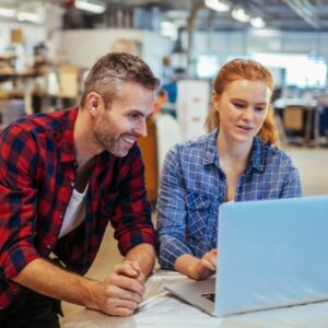 un hombre de mediana edad sonriente y una mujer joven en un taller comercial miran una computadora portátil